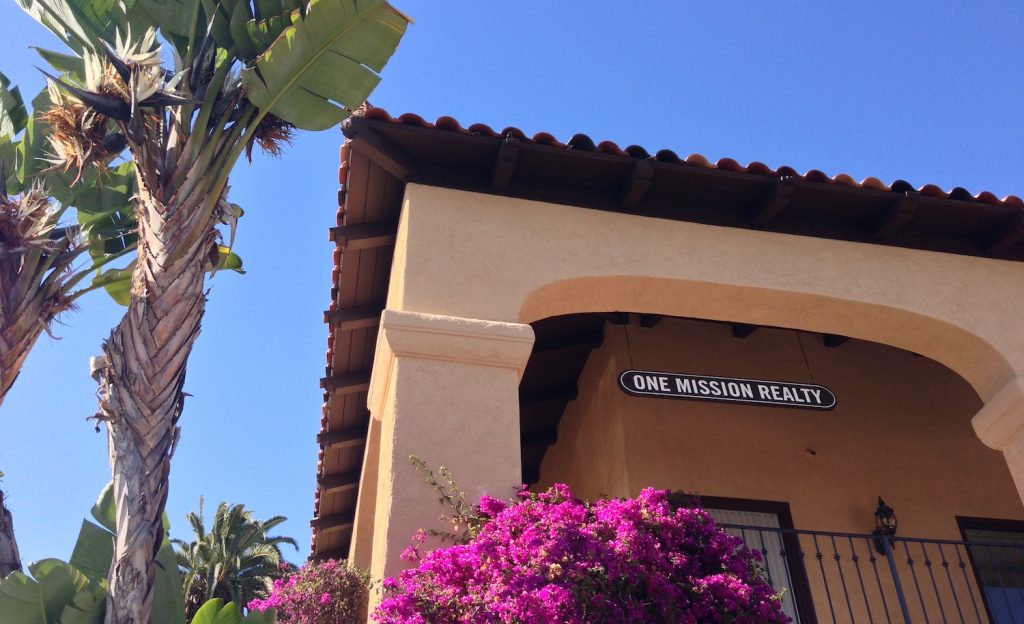 Spanish style building with a wooden sign hanging in an arch that reads "ONE MISSION REALTY." A large bird of paradise plant and a bougainvillea plant with pink flowers is present in the lower and left hand sides of the image. 