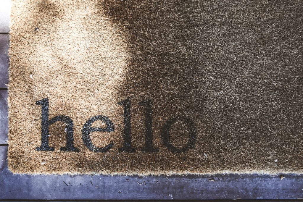 brown natural fiber door mat with the word 'hello' in all lower case lettering in the bottom left corner. Mat is set on a purplish-gray wood-slat floor.